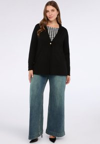 Žena oblečená v černém propínacím kardiganu přes top s černo-bílým houndstooth vzorem, širokých modrých džínech a černých špičatých lodičkách, stojící proti bílému pozadí.