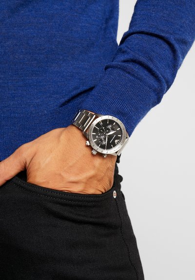 Polso con orologio cronografo in metallo argento, mano nella tasca dei pantaloni neri, indossa un maglione blu a maniche lunghe.