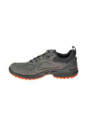 Grijze sportieve schoen met een gestructureerde suède en mesh bovenkant, voorzien van een zwarte kraag en een oranje rubberen zool voor grip. Vetersluiting.