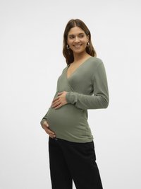 Porodniški top iz elastičnega olično zelene tkanine z V-izrezom in dolgimi rokavi, v kombinaciji s črnimi hlačami, ki poudarja okroglo nosečniško trebušček.