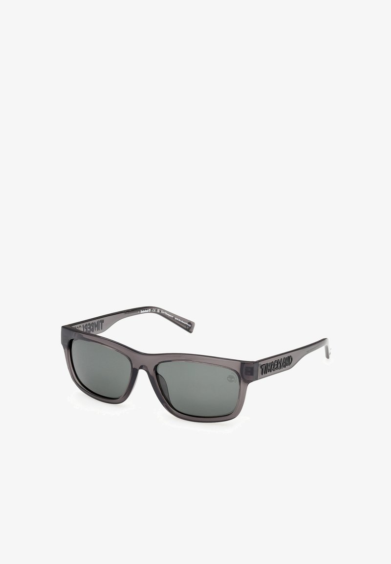Lunettes de soleil Timberland rectangulaires noires avec des verres teintés foncés et le nom de la marque sur les branches, présentées sur un fond blanc.