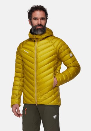 Veste isolante jaune avec un design en chevron, dotée d'une capuche et de poches zippées, associée à un pantalon sombre portant un logo.