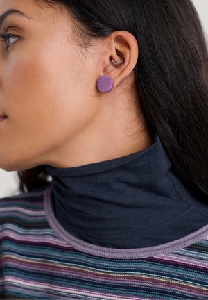 Orecchino a bottone rotondo in ceramica viola indossato all'orecchio, abbinato a un dolcevita scuro e a una maglietta a righe con vari colori.