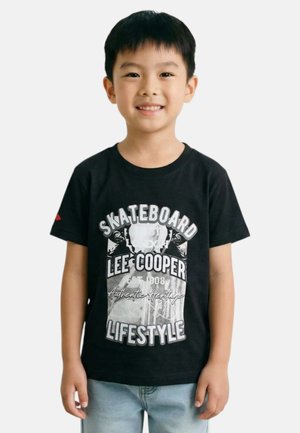 Jeune garçon souriant portant un T-shirt graphique noir avec le texte "Skateboard Lee Cooper Lifestyle" et un jean bleu clair, debout devant un fond blanc.