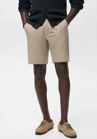Pantalones cortos beige de tela, diseño de frente plano, hasta la rodilla, combinados con zapatos de gamuza marrón claro y un cárdigan negro.