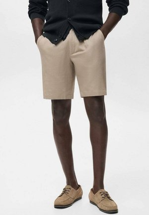 Pantalones cortos beige de tela, diseño de frente plano, hasta la rodilla, combinados con zapatos de gamuza marrón claro y un cárdigan negro.