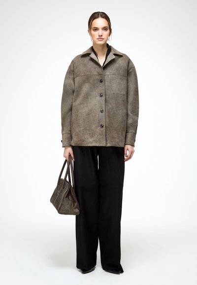 Žena v nadrozmernej hnedej semišovej bunde, čiernych širokých nohaviciach, drží hnedú textúrovanú kabelku na zápästí, stojí na bielom pozadí.