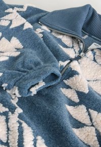 Sininen fleece-pusero ribatulla kauluksella, jossa on kermavärinen geometrinen kuvio. Sisältää vetoketju sulkemisen ja pehmeän, textuurisen kankaan.