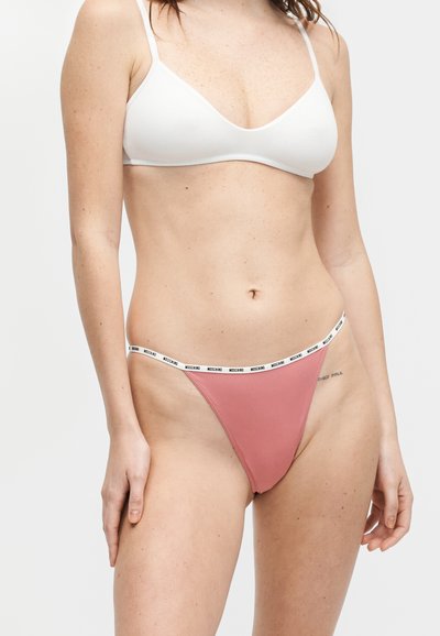 Femme portant un bralette blanc et un bas de bikini rose avec une ceinture blanche au logo Moschino, debout devant un fond uni.