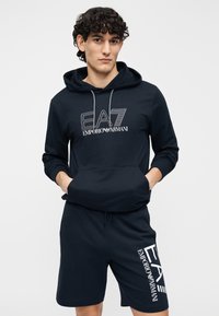 Mörkblå hoodie med framficka och dragsnören, med en vit grafisk logotyp. Matchas med matchande shorts med logotyp på sidan.