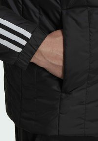 Zwarte pufferjack met een hoge kraag, met een zichtbare hand in een zijzak en witte gestreepte accenten op de mouw. Gestructureerde stof.