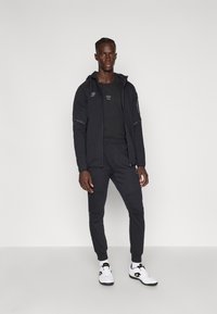 Čierny športový outfit pozostávajúci z mikiny na zips s detailmi loga, zúžených nohavíc a bielych tenisiek s čiernymi akcentmi.