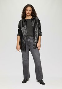 Schwarze Kunstleder-Hemdjacke über einem grauen Pullover, kombiniert mit grauen weit geschnittenen Jeans und schwarzen Stiefeletten. Strukturierte Materialien, lässige Passform.
