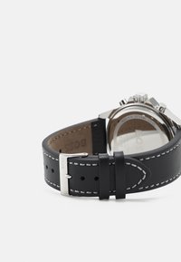 Correa de reloj de cuero negra con costuras blancas, herrajes metálicos plateados y un detalle de logo. Caja redonda con fondo transparente.