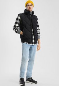 Schwarze gesteppte Weste mit hohem Kragen, getragen über einem schwarz-weiß karierten Hemd, hellblauen Jeans und schwarzen Sportschuhen. Gelbe Strickmütze.