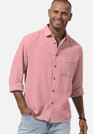 Man smiling wearing a long-sleeve pink textured button-up shirt with rolled cuffs and light blue jeans, hand in pocket.