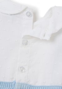 Blusa de tela blanca con pequeños detalles texturizados. Presenta un cuello redondeado, botones transparentes y un dobladillo de punto en azul claro.