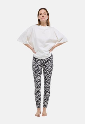 Femme debout pieds nus, mains sur les hanches, portant un haut blanc ample à manches courtes et des leggings moulants à motif léopard sur un fond uni.