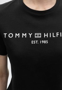 Μαύρο t-shirt με λευκό κείμενο "Tommy Hilfiger" και λογότυπο, με την ένδειξη "EST. 1985" τυπωμένη κάτω από το όνομα της μάρκας στην περιοχή του στήθους.