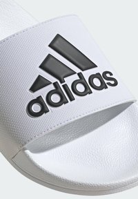 Infradito bianchi con tomaia testurizzata, dotati di un prominente logo Adidas nero sulla fascia. Suola piatta con una finitura liscia.