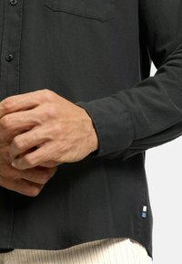 Black button-up shirt with a herringbone pattern, featuring a chest pocket and a small colored tag at the hem. Long sleeves are cuffed.