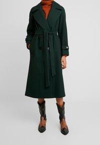 Tummanvihreä pitkä trenssitakki, jossa vyötäröllä solmittava vyö, leveä kaulus ja kaksi etutaskua. Yhdistetty mustiin ja ruskeisiin cowboy-saappaisiin, joissa on tähtiä korostuksena.