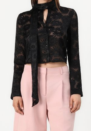 Czarna transparentna bluzka z kwiatowym wzorem i długim wiązaniem na szyi, zestawiona z jasnoróżowymi spodniami z wysokim stanem na stojącej osobie.