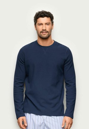 Mann mit dunklem lockigem Haar und Bart, der ein marineblaues Langarmhemd und helle gestreifte Hose trägt, steht vor einem einfarbigen Hintergrund.