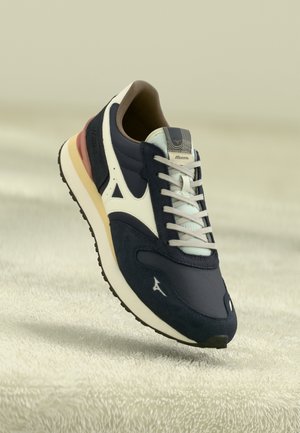 Granatowo-białe buty sportowe Mizuno z beżowymi i rdzaawymi akcentami, białymi sznurówkami i teksturowaną czarną podeszwą na jasnej wykładzinie.