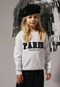Valge särk, millel on mustad litrid ja kiri "PARIS", ning musta skripti tipp "Mentally In Paris". Must litritega barett, pikk blond juuksed, neutraalne taust.