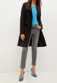 Svart kappa, blå topp, grå jeans med sidmönster och blå höga klackar. Kappan är skräddarsydd; jeansen har en slim fit.