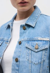 Φωτεινό γαλάζιο τζιν μπουφάν με κλασικό γιακά, δύο τσέπες στο στήθος, χάλκινα κουμπιά και ετικέτα που αναγράφει "Pepe Jeans London." Λευκό μπλουζάκι από κάτω.