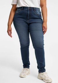 Mörkblå högmidjade jeans med en slät yta, fem fickor och kontrasterande orange sömmar, kombinerat med ljusa sneakers.