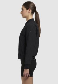 Giacca bomber nera trapuntata con polsini e colletto a coste. Caratterizzata da una cerniera e cuciture decorative. Vista laterale.