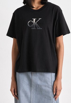 Personne portant un t-shirt noir Calvin Klein Jeans avec un grand logo "CK", assorti à une jupe en denim bleu clair.