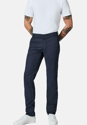Mann mit Tattoos, der dunkelblaue Hose, weißes kurzärmeliges Hemd und weiße Sneaker trägt, steht mit den Händen hinter dem Rücken.