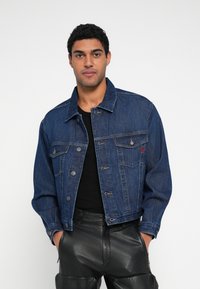 Τζιν μπουφάν σε σκούρο μπλε με κλασική γραμμή, τσέπες μπροστά, μεταλλικά κουμπιά και λεπτή κόκκινη λεπτομέρεια στο αριστερό στήθος. Μαύρο πουκάμισο από κάτω.