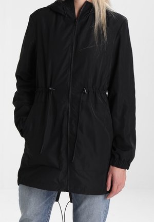Person trägt eine schwarze Kapuzenjacke mit Reißverschluss, elastischen Bündchen und Kordelzug an der Taille, kombiniert mit hellblauen Jeans.