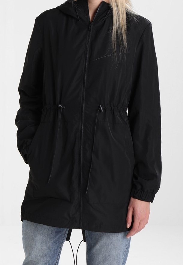 Personne portant une veste à capuche noire zippée avec des poignets élastiques et des cordons à la taille, associée à un jean bleu clair.