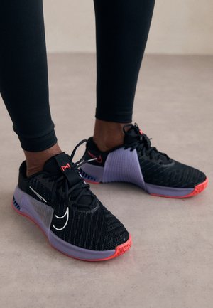 Person trägt schwarze und violette Sportsneaker mit roten Sohlen, steht auf einem hellen Boden und trägt schwarze Leggings.