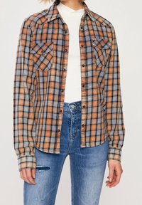 Camicia a quadri nei toni dell'arancione e del blu, con due tasche sul petto, chiusura con bottoni e maniche lunghe. Indossata sopra a un top bianco.