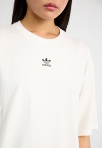 Bílý bavlněný tričko s krátkými rukávy, s malým černým logem Adidas na hrudi. Hladká textura a pohodlný střih.