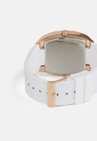 Montre avec un bracelet en caoutchouc blanc, boîtier carré doré, bande texturée et fermoir à bouton-poussoir. L'arrière présente des détails gravés et une finition mate.