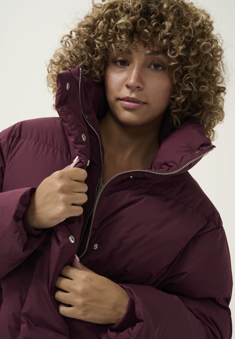 Bordeaux pufferjack met hoge kraag, zilveren ritssluiting en drukknoopsluiting, met een gemêleerde stof en geribbelde manchetten.
