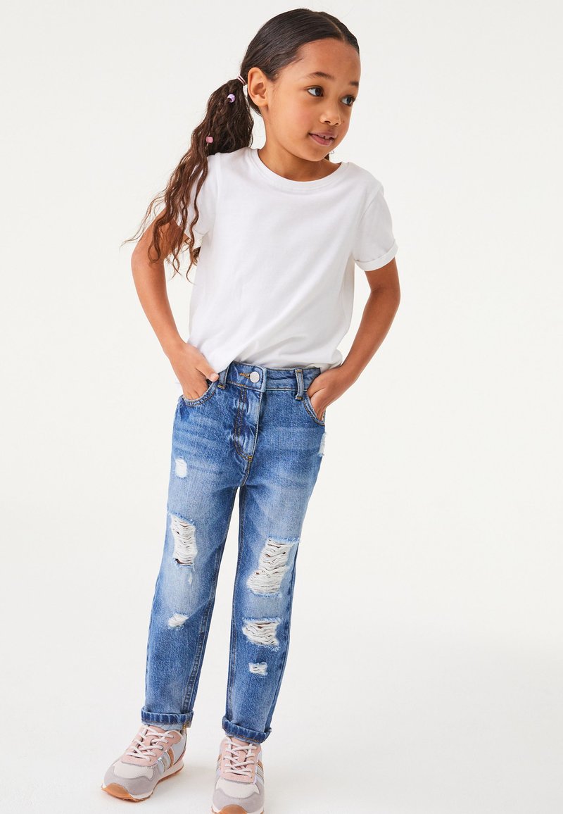 Jeans blu strappati con strappi, abbinati a una maglietta bianca. Il bambino indossa sneakers rosa chiaro e grigie. Lo sfondo è bianco uniforme.
