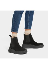 Botines de ante negros con suela de goma, paneles laterales elásticos y una pestaña para tirar. Llevados con jeans azules y calcetines beige.