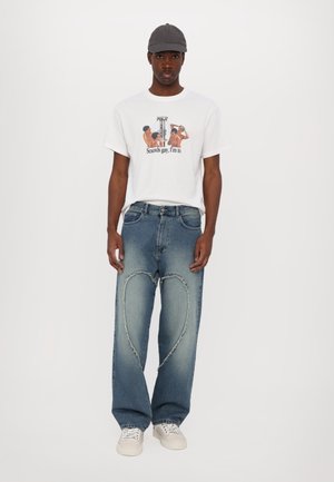 Witte t-shirt met korte mouwen en grafische print; blauwe spijkerbroek met hartvormige uitsparingen; witte sneakers; grijze pet.