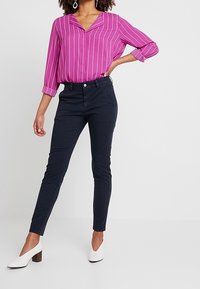 Różowo-biała bluzka w paski z dekoltem w szpic, zestawiona z dopasowanymi ciemnogranatowymi spodniami. Modelka nosi białe buty na obcasie typu slip-on, stoi na białym tle.