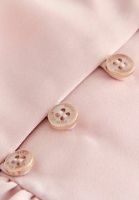 Helles rosa Material mit einer glatten Textur verfügt über drei Knöpfe. Die Knöpfe sind cremefarben mit eingraviertem Text und entlang der Stoffnaht angeordnet.