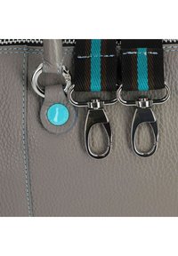 Detalle de bolso de cuero gris con herrajes plateados, una etiqueta de acento turquesa y una superficie texturizada con costuras azules. Se ven dos ganchos de presión.